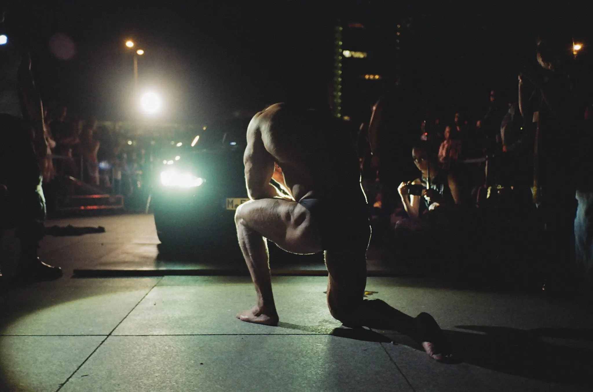 A bodybuilder performes for Kunak. He is kneeled in front of a car in the night.