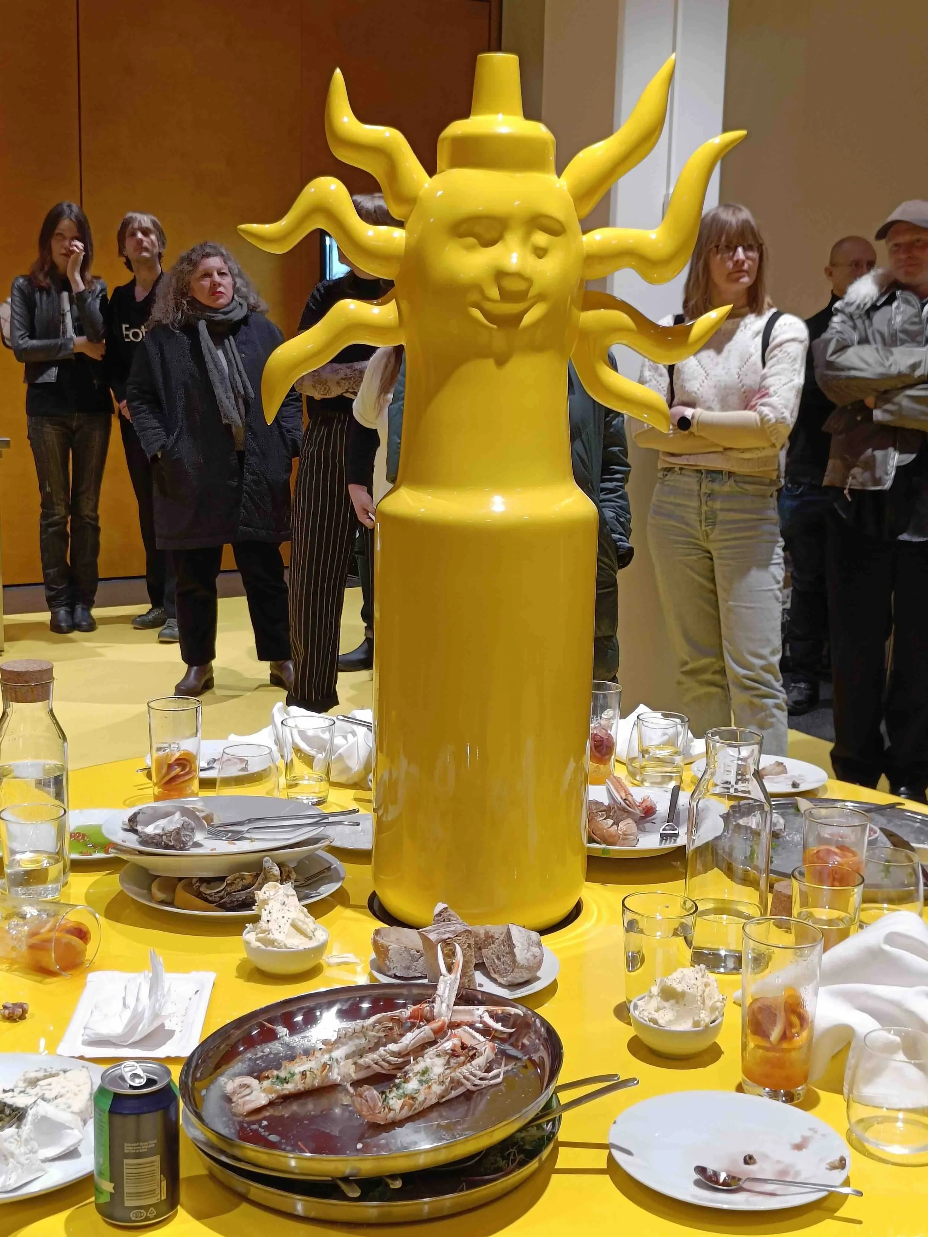 A bright yellow sun table is covered with empty food spectators are surrounding it