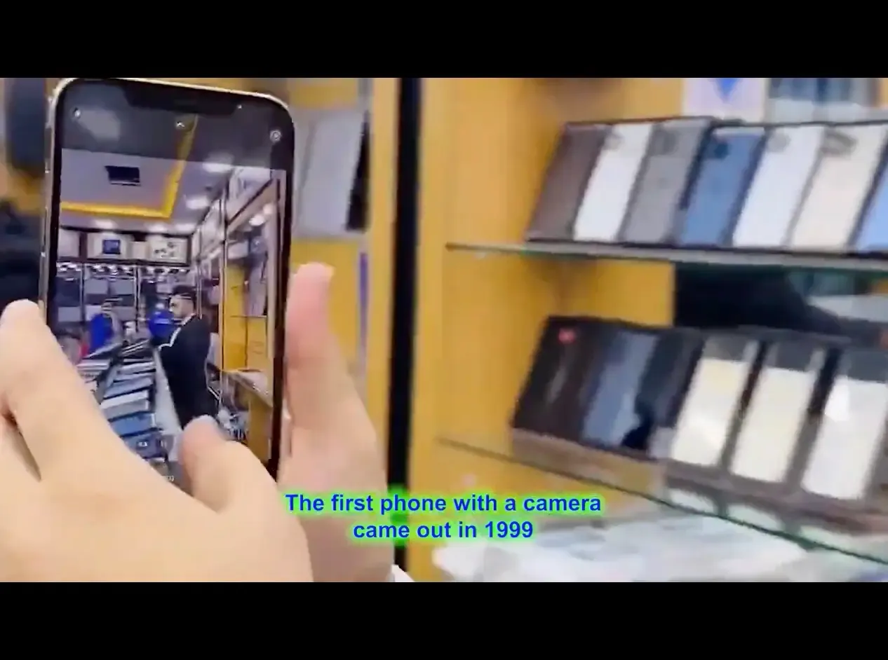 A caption of an iphone in a phone shop. Subtitles read "The first phone with a camera came out in 1999"
