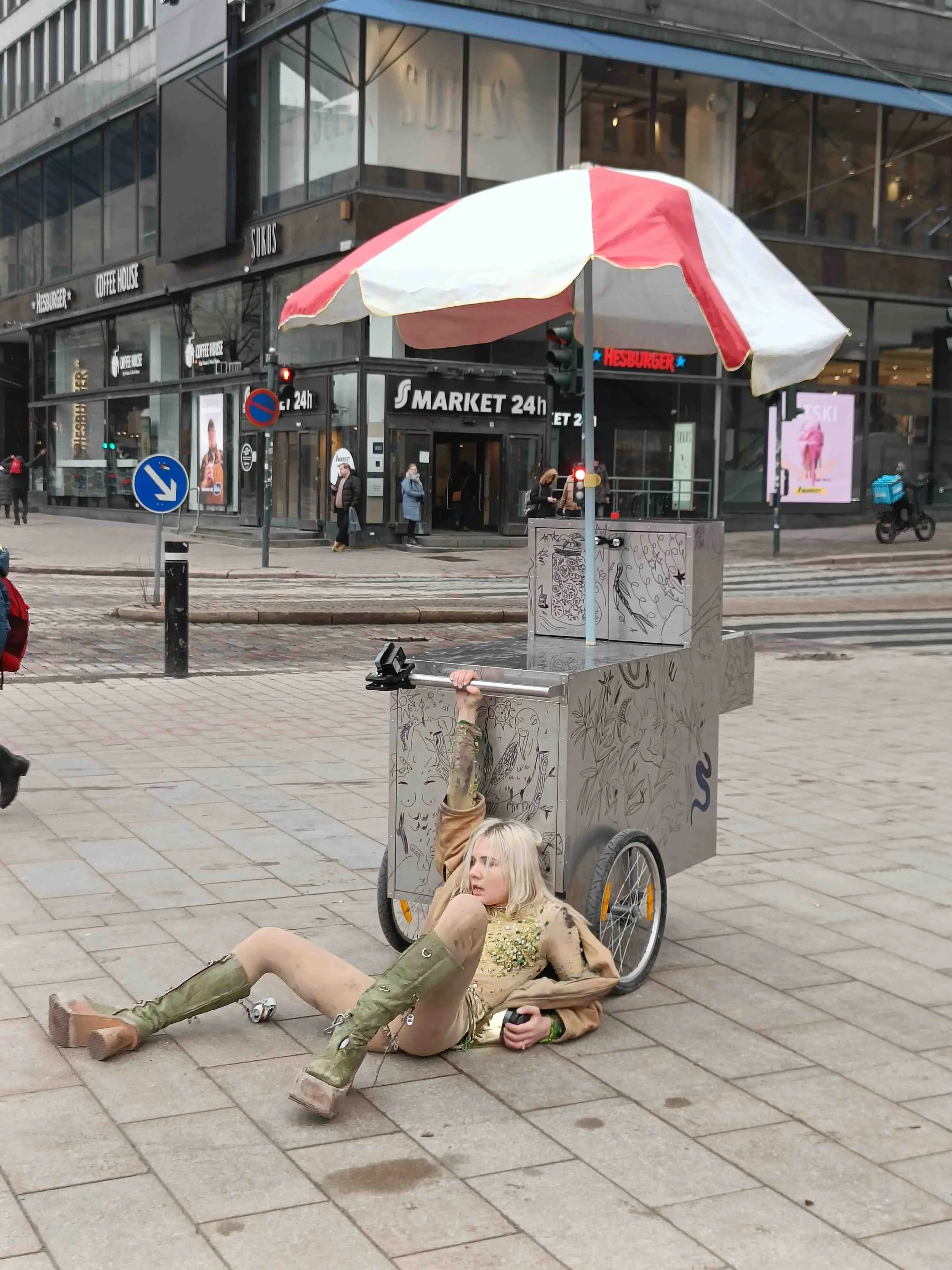 Maria Metsalu is laying on the floor holding a hot dog chart, performing outside in the street